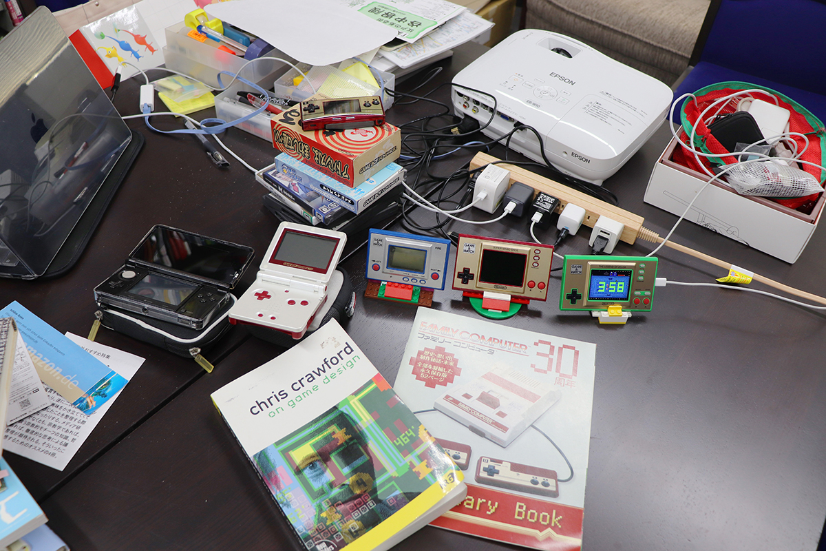 研究室の机の上に「ゲーム&ウオッチ」「ゲームボーイ」などのデバイスや関連図書が置かれている